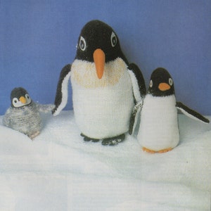 May include: Three knitted penguin toys, one large, one medium, and one small. The penguins are white with black heads and beaks, and orange beaks. The penguins are sitting on a white surface that looks like snow.