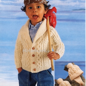 Może przedstawiać: Małe dziecko w białym dzianym kardiganie z wzorem warkocza i brązowymi guzikami. Kardigan ma kołnierz szalowy. Dziecko ma na sobie również niebieskie dżinsy i brązową chustkę z białym wzorem. Dziecko trzyma drewniany kij. W prawym dolnym rogu zdjęcia widać dwa białe dziane czapki z pomponami.