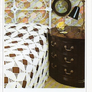 Peut inclure: Intérieur de chambre avec un lit en fer forgé blanc et une table de chevet en bois foncé. Le lit est recouvert d'une couverture blanche à tissage ouvert. Une petite image encadrée et une lampe sont sur la table. Le papier peint a un motif patchwork.