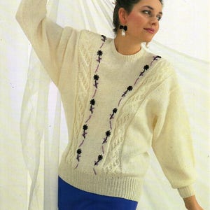 Könnte beinhalten: Ein weißer Strickpullover mit Zopfmuster und schwarzer und violetter Blumenstickerei. Der Pullover hat lange Ärmel und einen Rundhalsausschnitt. Der Pullover ist 76-102 cm (30-40 Zoll) groß und in sechs Größen erhältlich.