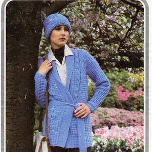 May include: A blue knitted cardigan with a cable knit pattern and a matching blue knitted hat. The cardigan has a wrap style and a belt. The model is wearing the cardigan and hat in a garden setting.