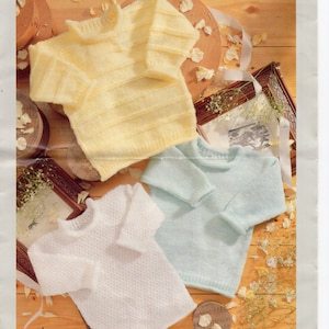 May include: Three baby sweaters in different colours: yellow, light blue and white. The sweaters are made with a double knit stitch and have a ribbed neckline and cuffs. The sweaters are laid out on a wooden surface with white flower petals scattered around them. The image includes the text "BABY DOUBLE KNITTING" and "SPECTRUM 7122 30-61cm/12-24ins".