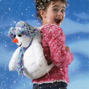 May include: A young girl wearing a pink fuzzy sweater and blue jeans is holding a white crocheted snowman with a purple and blue hat and scarf. The girl is smiling and the background is a blue sky with white snowflakes.