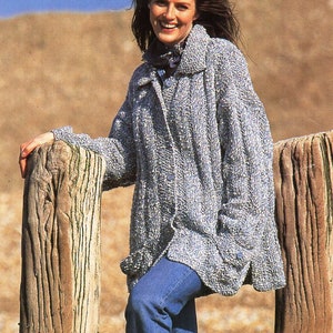 May include: A woman wearing a blue and white textured knit coat with a collar and button closure, paired with blue jeans. She is leaning against a weathered wooden fence, smiling. The background features a sandy beach and blue sky.