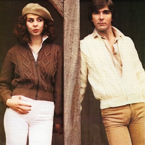 May include: A woman wearing a brown sweater and a beige beret stands next to a man wearing a white sweater and brown pants. They are both standing next to a wooden post.