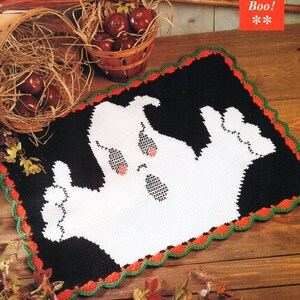 May include: A crocheted Halloween doormat with a white ghost design on a black background. The border is a scalloped edge with green and orange trim. The text "13 Boo!" is in the upper right corner.