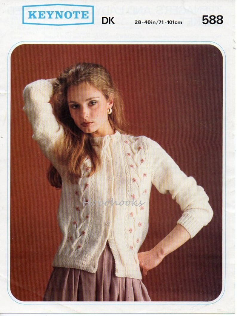 K&ouml;nnte beinhalten: Ein wei&szlig;er Strickcardigan mit einem Zopfmuster und rosa Blumen Stickerei. Der Cardigan hat einen Knopfverschluss und wird an einem Model mit einem braunen Rock gezeigt. Die Musternummer ist 588.