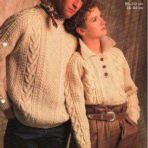 Puede incluir: Dos jerséis de punto Aran de color crema, uno con cuello redondo y el otro con cuello y botones. Los jerséis están hechos con un patrón de punto de cable. Los jerséis son usados por dos personas, una de pie detrás de la otra. El texto "Sunbeam Aran 1158 86-112 cm 34-44 ins" está impreso en la imagen.
