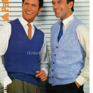 May include: Two men wearing white shirts and knitted vests. The vest on the left is blue and the vest on the right is light blue. Both vests have a V-neck and are buttoned up the front. The men are standing in front of a white slatted blind.