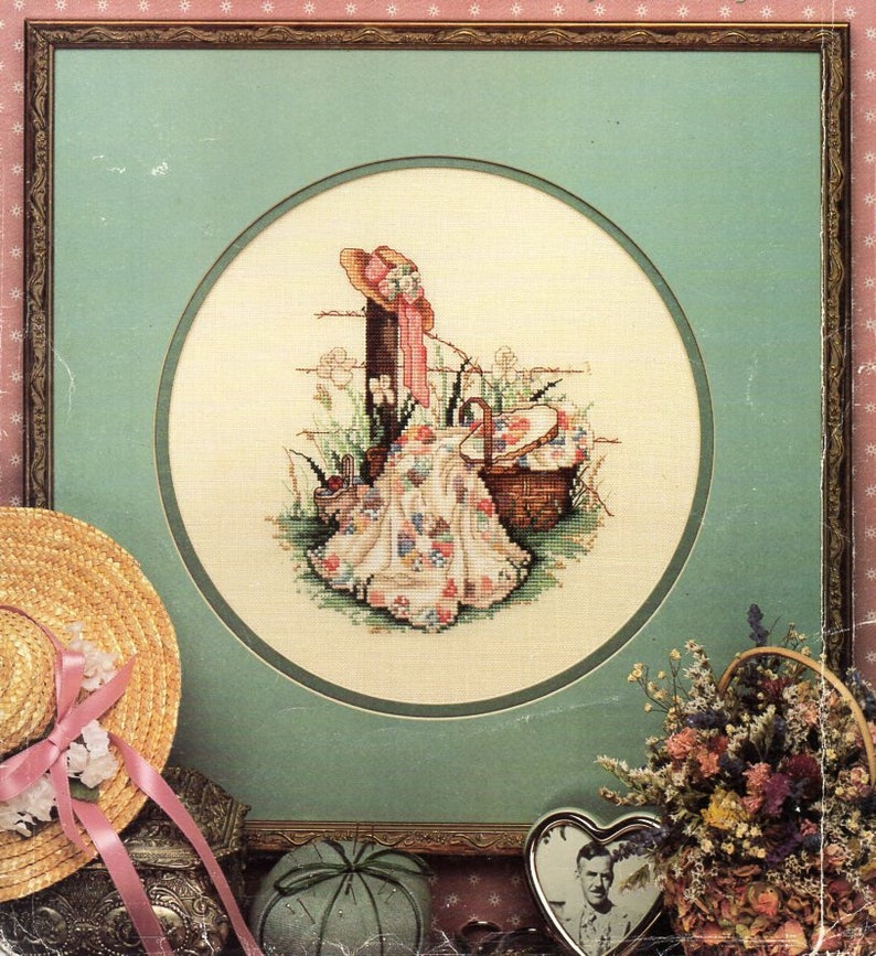 May include: A framed cross-stitch embroidery depicting a woman in a pink hat and dress sitting on a blanket with a picnic basket. The embroidery is surrounded by a green mat and a gold frame.