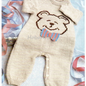 May include: A knitted baby onesie in a light beige color with a brown bear face and a striped bow tie. The onesie has a button closure on the crotch.