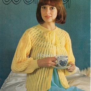 Könnte beinhalten: Ein gelber Strickcardigan mit einem Spitzenmuster und einer Schleife. Der Cardigan wird von einer Frau mit kurzen braunen Haaren getragen. Die Frau hält eine Teetasse und eine Untertasse.