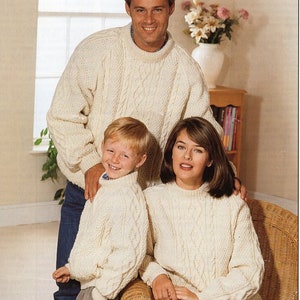 Puede incluir: Una familia de tres personas con suéteres de punto blanco con cable. El padre está de pie con el brazo alrededor de su hijo, que está de pie frente a él. La madre está sentada en una silla de mimbre con el brazo alrededor de la cintura del padre.