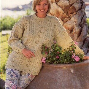 May include: A pale green cable-knit jumper with a round neck and long sleeves. The jumper has a textured design and is worn with floral leggings. The person is leaning against a large, brown terracotta pot with pink flowers.