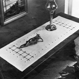 May include: A white table runner with a black and white embroidered design. The runner is on a wooden table with a silver figurine of a fox and a glass lamp.