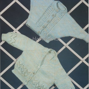 May include: Two knitted baby cardigans, one light blue and one light green, with a button closure and lace detailing. The cardigans are hanging on a white lattice background.