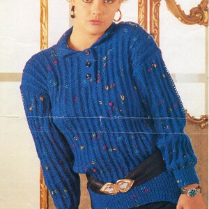Può includere: Un maglione blu lavorato a maglia con collo e tre bottoni. Il maglione ha una trama a coste ed è decorato con macchie colorate. Una cintura nera con fibbia dorata stringe la vita. L'immagine mostra anche un orologio con cinturino argentato.