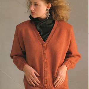 May include: A rust-coloured knitted cardigan with a V-neck and button fastening. The cardigan has long sleeves and a pocket on the left side. The model is wearing a black turtleneck underneath.