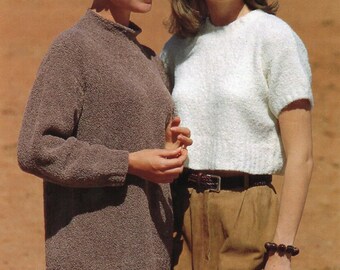 Op de afbeelding: Twee vrouwen dragen truien. De vrouw links draagt een bruine trui en een witte broek. De vrouw rechts draagt een witte trui en een bruine broek met een bruine riem.