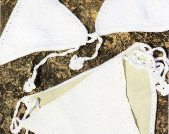 May include: A white, handmade crochet bikini set. The top is a triangle style with tie closures, and the bottoms are a classic cut. The bikini is displayed on a textured, brown surface.