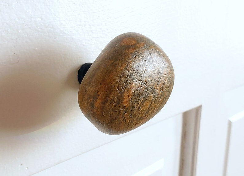 Może przedstawiać: Gałka do szafki z naturalnego kamienia w odcieniach brązu i beżu. Owalna gałka zamontowana na białych drzwiach szafki. Unikalny, rustykalny akcent.