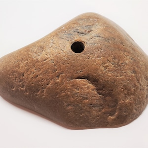May include: A brown, irregularly shaped stone with a hole drilled through the center. The stone has a rough, textured surface.