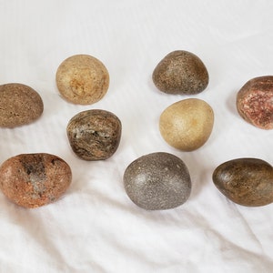 Może przedstawiać: Kolekcja dziewięciu gładkich, zaokrąglonych kamieni w r&oacute;żnych odcieniach ziemi, od beżu po czerwonobrązowy. Kamienie, o r&oacute;żnych rozmiarach i kształtach, ułożone są na białej powierzchni. Niekt&oacute;re mają średnicę od 3 cm do 6 cm.