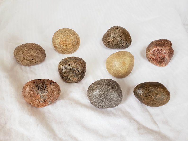 Może przedstawiać: Kolekcja dziewięciu gładkich, zaokrąglonych kamieni w r&oacute;żnych odcieniach ziemi, od beżu po czerwonobrązowy. Kamienie, o r&oacute;żnych rozmiarach i kształtach, ułożone są na białej powierzchni. Niekt&oacute;re mają średnicę od 3 cm do 6 cm.