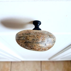 Może przedstawiać: Unikalny uchwyt do szafki z uchwytem z naturalnego kamienia i czarną metalową podstawą. Kamień to mieszanka beżu, brązu i szarości, o gładkim, zaokrąglonym kształcie. Ten dekoracyjny element dodaje rustykalnego charakteru.