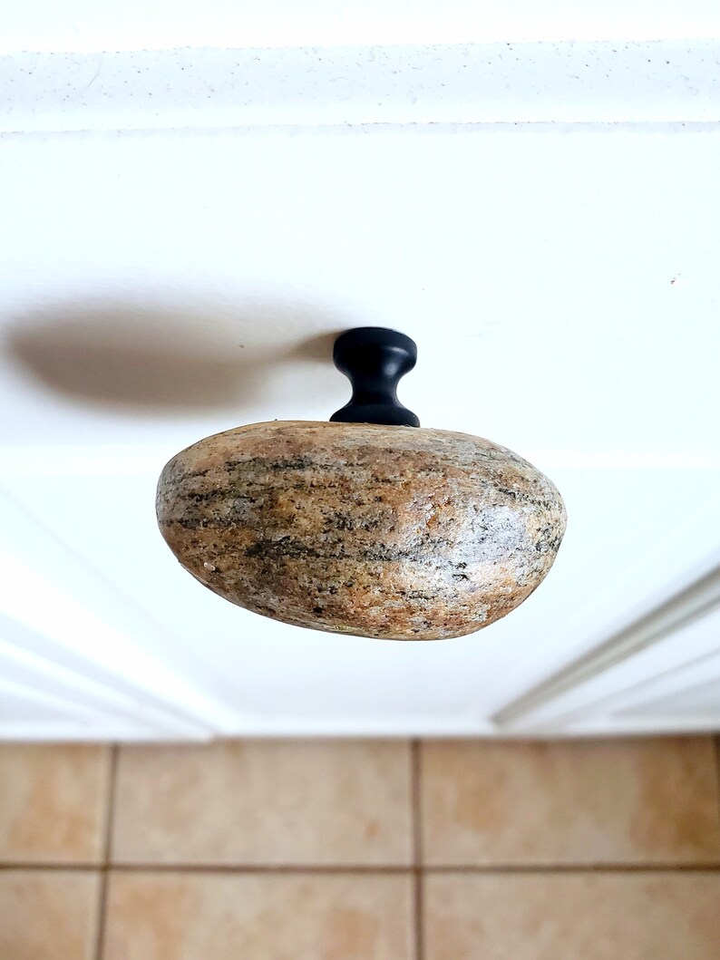 Może przedstawiać: Unikalny uchwyt do szafki z uchwytem z naturalnego kamienia i czarną metalową podstawą. Kamień to mieszanka beżu, brązu i szarości, o gładkim, zaokrąglonym kształcie. Ten dekoracyjny element dodaje rustykalnego charakteru.