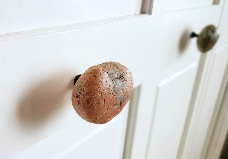 Może przedstawiać: Zbliżenie białych drzwi z dwoma kamiennymi gałkami. Gałki mają owalny kształt, z mieszanką brązowych, szarych i beżowych kolor&oacute;w. Drzwi mają prosty, klasyczny design.