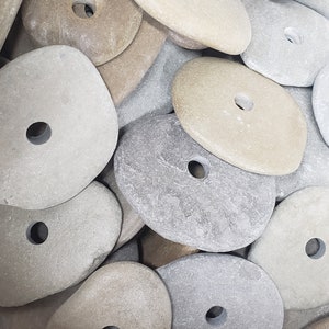 May include: A collection of flat, smooth, rounded stones in shades of tan, beige, and gray. Each stone features a single, centered, circular hole drilled through the middle, creating a uniform appearance across the varied natural rock shapes.