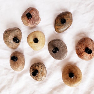 Może przedstawiać: Widok z g&oacute;ry na dziewięć naturalnych kamieni rzecznych, każdy z czarnym, cylindrycznym przedmiotem włożonym w wywiercony otw&oacute;r. Kamienie mają r&oacute;żne kolory, od beżowego do czerwonobrązowego, na białej, teksturowanej powierzchni.