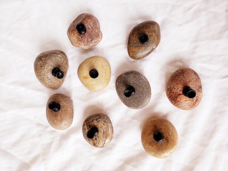 Może przedstawiać: Widok z g&oacute;ry na dziewięć naturalnych kamieni rzecznych, każdy z czarnym, cylindrycznym przedmiotem włożonym w wywiercony otw&oacute;r. Kamienie mają r&oacute;żne kolory, od beżowego do czerwonobrązowego, na białej, teksturowanej powierzchni.