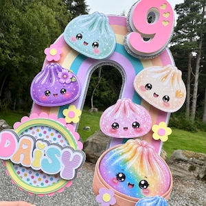 May include: A layered cake topper featuring a large rainbow arch, the number 9, and the name DAISY in pastel letters. The design includes several cute, smiling dumpling characters with glittery details, small paper flowers, and a bamboo steamer base.