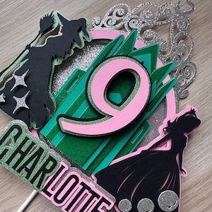 May include: A pink and green cake topper featuring a silhouette of a witch, a castle, and a princess. The number 9 is in the centre of the castle. The name Charlotte is written in pink and black letters.