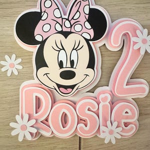 May include: A layered cake topper featuring Minnie Mouse with a pink bow and the number 2. The name "Rosie" is also displayed in pink, with white daisy accents. The design is set against a light wood grain background.