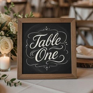 May include: A wooden-framed chalkboard sign with the words "Table One" in elegant white script. The sign sits on a white tablecloth, next to a floral arrangement of white roses and lit candles, suggesting a wedding or event setting.
