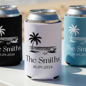 May include: Three beverage can coolers in black, white, and blue, each with a palm tree, seashell, and the text "The Smiths" and the date 10.09.2024. The coolers are on a white table with a beach and ocean background.