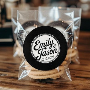 May include: Clear cellophane bags filled with cookies, each bag sealed with a black circular sticker. The sticker features the names "Emily & Jason" and the date "12.10.2026" in white script. The cookies are light brown and stacked.