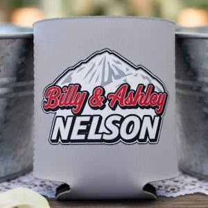 May include: A gray can cooler with the names "Billy & Ashley" in red script and "NELSON" in white block letters, beneath a mountain graphic. The cooler is positioned between two galvanized metal buckets.