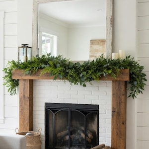 May include: A white brick fireplace with a black metal screen, flanked by wooden posts. A mirror hangs above the mantel, adorned with greenery and candles. A woven basket and firewood add to the cozy aesthetic.
