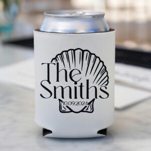 May include: White can cooler with a black seashell design and the text "The Smiths" with the date "10.09.2024". The cooler is designed to keep beverages cold and is made of a soft, insulating material.