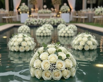 May include: Multiple spherical floral arrangements consisting of white roses and small white baby's breath flowers floating on the surface of a dark pool, with a blurred outdoor event setting featuring white curtains and lights in the background.