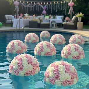May include: Floating floral arrangements in a pool. Each arrangement is a dome shape, composed of pink and white artificial roses. The water is a clear blue, and the scene suggests a party or event with decorations in the background.