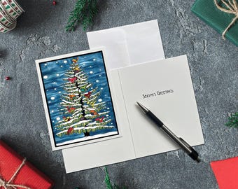 May include: A Christmas card with a watercolor illustration of a decorated Christmas tree, with the words "Season's Greetings" inside. A pen rests on the open card. The card is on a gray surface with holiday decorations.