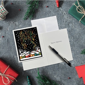 May include: A holiday greeting card with a black and white illustration of a snowy village with colorful houses. The card is open, revealing the text "Season's Greetings" and a pen. The scene is set on a gray surface with festive decorations.