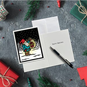 May include: A greeting card with a black border features a cartoon gnome holding a lantern in a snowy scene. The card is open, revealing the message "Season's Greetings" and a pen. The scene is set on a gray surface with holiday decorations.