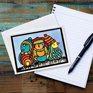 May include: A greeting card featuring three colorful cartoon birds wearing winter hats and scarves. The birds are set against a light blue background, with a black border. The card is on a white envelope, with a lined notepad and pen.