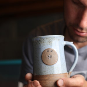 May include: A blue and brown ceramic mug with a brown circle on the side that has the letters "OX" and a star inside. The mug has a handle.